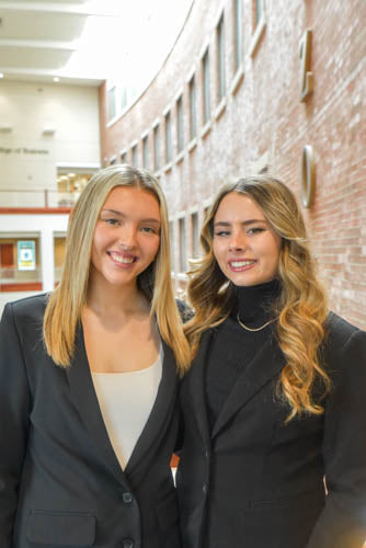 Two women, Elizabeth (Lizzy) Dekrell and Alyssa Pick in formal attire standing together in a hallway for a professional headshot promoting their gluten-free snack box brand.
