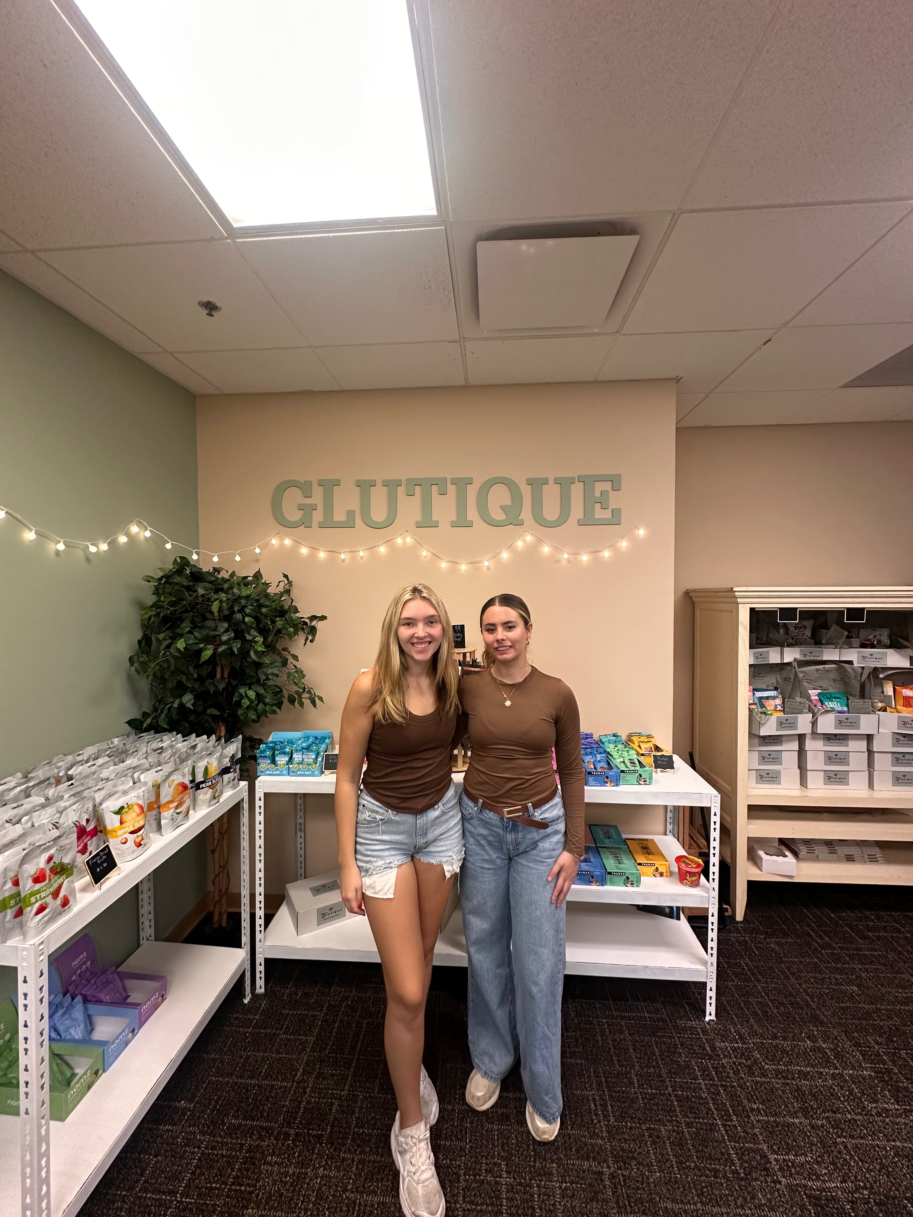 Glutique cofounders Elizabeth and Alyssa standing inside their gluten-free snack shop with the Glutique logo displayed on the wall.