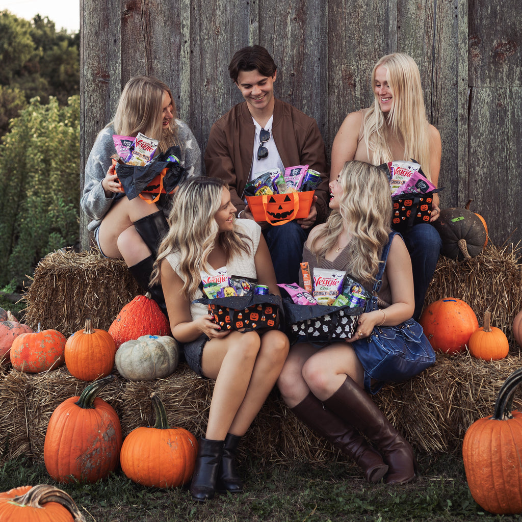 Group of friends sitting on hay bales holding Glutique Halloween Boo Boxes filled with gluten-free snacks, with pumpkins in the background during a fall photo shoot.