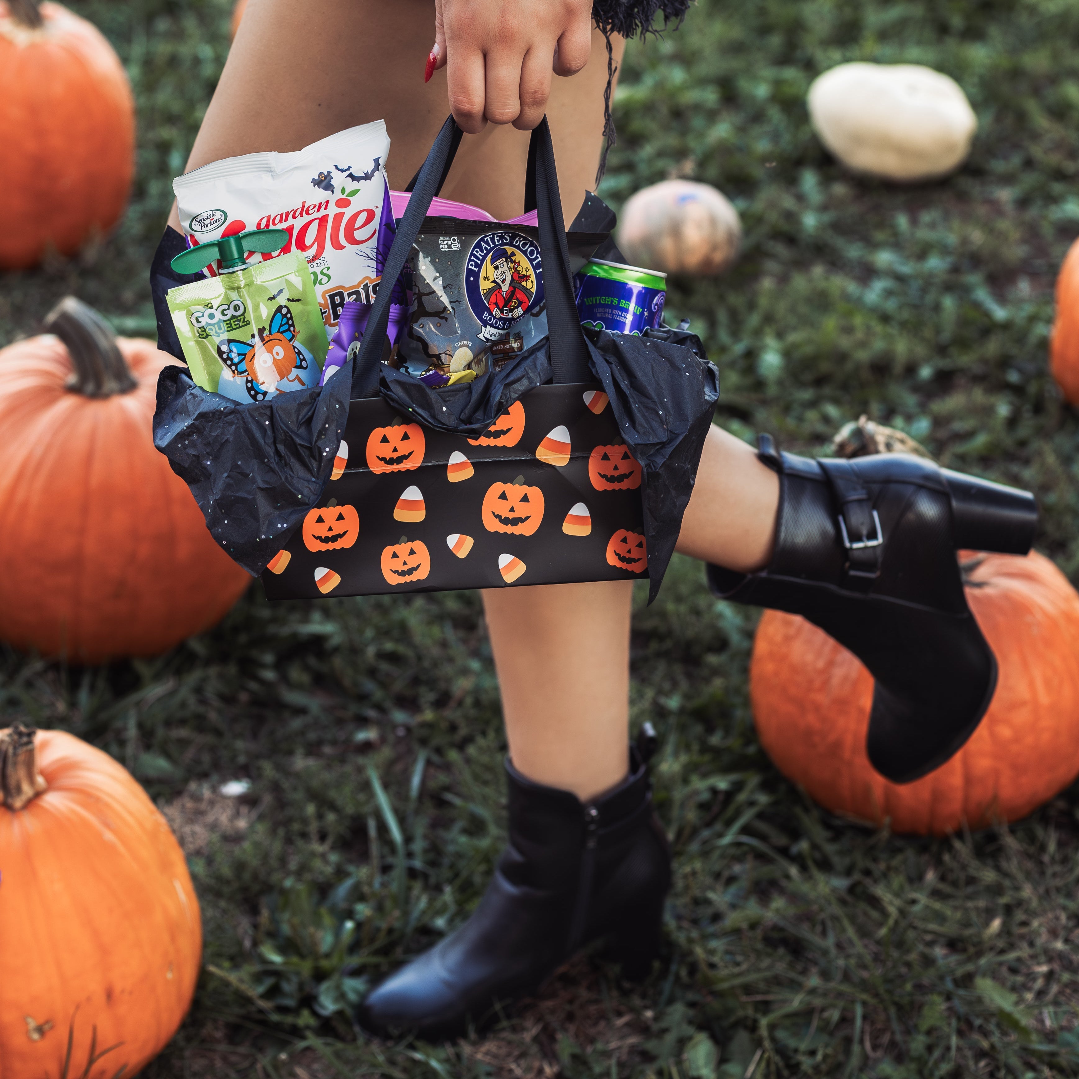Girl walking through a pumpkin patch holding a Glutique Halloween Boo Box filled with gluten-free snacks, candy, and drinks. Alani Witches Brew