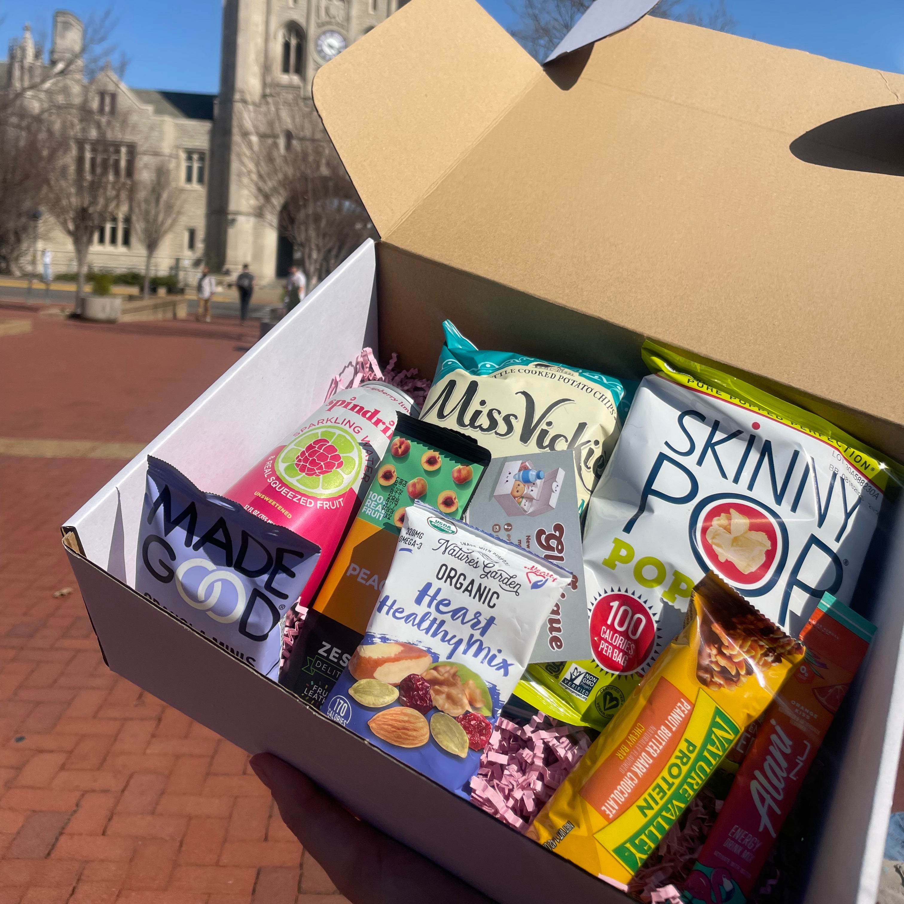 Open Glutique gluten-free snack box filled with healthy snacks like Skinny Pop, MadeGood, and Miss Vickie’s, displayed on a college campus